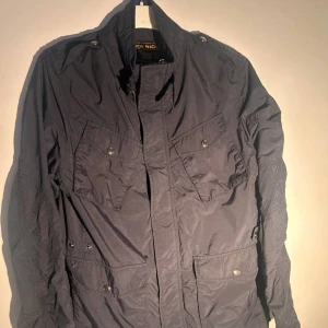 Woolrich windbreaker - Snygg svart vindjacka från Woolrich med flera fickor framtill och coola detaljer som axelklaffar och dragkedja. Jackan har en klassisk ståkrage och är tillverkad i lätt polyester, perfekt för blåsiga dagar. Insidan är fodrad i blått för extra stilpoäng.