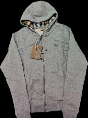 Grå zip-hoodie från Burberry - Snygg grå zip-hoodie från Burberry med klassiskt rutigt foder i huvan och diskret logga på bröstet. Hoodien har dragsko i huvan, fickor fram och ribbade muddar. Perfekt för en chill och stilren look. Aldrig använd 