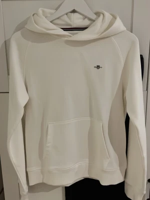 Vit hoodie från Gant - Vit hoodie från Gant med broderad logga på bröstet. Klassisk modell med huva och känguruficka framtill. Mjuk bomullskänsla och stilren design som passar perfekt till jeans eller mjukisbyxor.
