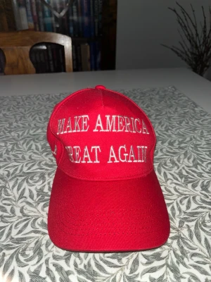 Röd keps Make America Great Again - Röd keps med böjd skärm och vit brodyr med texten 'MAKE AMERICA GREAT AGAIN' framtill och '45-47' på sidan. Klassisk basebollmodell med justerbar passform. Perfekt för dig som vill sticka ut med en ikonisk accessoar.