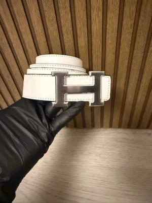 Vitt bälte med H-spänne - Snyggt vitt bälte med det ikoniska H-spännet i silverfärgad metall. Perfekt accessoar för att lyfta din outfit med en exklusiv touch. Klassisk design och stilren look som passar till mycket.