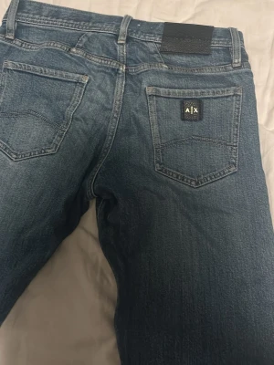 Blå jeans från Armani Exchange size 29 - Snygga blå jeans från Armani Exchange med klassisk femficksdesign och A|X-logga på bakfickan. Jeansen har rak passform och är tillverkade i mjukt denimtyg som ger en avslappnad look. Perfekta till en casual outfit.
