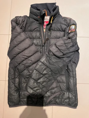 Svart dunjacka Parajumpers herr - Svart dunjacka från Parajumpers med quiltad design och hög krage. Jackan har dragkedja framtill, två sidofickor med dragkedja och patch med logga på ena ärmen. Insidan är ljusgrå och jackan är tillverkad i syntetmaterial med dunfyllning. Perfekt för kalla dagar. Jackan passar på dig som är omkring 160-170.
