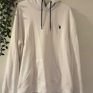 Vit unisex hoodie från U.S. Polo Assn. Storlek XL - Snygg vit hoodie från U.S. Polo Assn. med svart broderad logga på bröstet och svarta dragsnören i huvan. Klassisk känguruficka framtill och mjukt material som känns skönt mot huden. Perfekt för en avslappnad och stilren look.