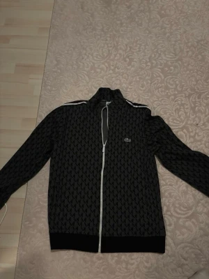 Svart ziptröja från Lacoste - Snygg svart ziptröja från Lacoste med vitt blixtlås och mönster i grått. Tröjan har ståkrage, vita ränder på axlarna och den klassiska gröna krokodilloggan på bröstet. Perfekt för en sportig och trendig look.