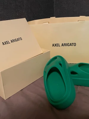 Gröna tofflor från Axel Arigato - Säljer ett par gröna tofflor från Axel Arigato. De har en chunky, modern design med rund tå och platt sula. Tillverkade i syntetmaterial och perfekta för dig som vill ha något stilrent och bekvämt att glida runt i. 