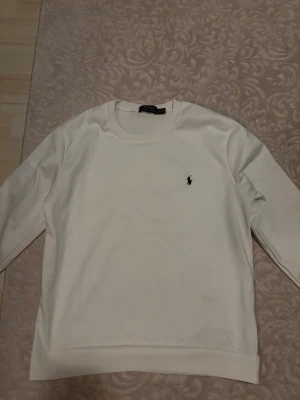 Vit sweatshirt från Polo Ralph Lauren - Klassisk vit sweatshirt från Polo Ralph Lauren med rund halsringning och den ikoniska broderade loggan på bröstet. Tröjan har långa ärmar och ribbade muddar vid ärmslut och nederkant. Perfekt för en clean och stilren look.