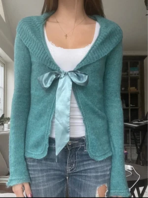 Turkos kofta med sidenrosett - Söt turkos kofta med lång ärm och stor krage. Koftan knyts framtill med en bred sidenrosett i matchande färg. Materialet ser mjukt och lite fluffigt ut, perfekt för lager på lager. Passar till jeans eller kjol för en trendig look. Vintage från Bik Bok💗