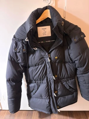 Svart dunjacka med huva från Boomerang  - Snygg svart dunjacka från Boomerang med avtagbar huva, dragkedja och knappar framtill. Jackan har två stora fickor, justerbar midja och detaljer i metall. Perfekt för kalla dagar och riktigt varm tack vare dunfyllningen.