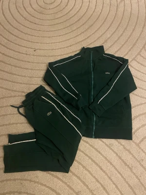 Grön set från lacoste - Mörkgrön set från Lacoste med hel dragkedja framtill och vita detaljer längs ärmarna. Klassisk hög krage och diskret Lacoste-logga på bröstet. Västen har en sportig look och är tillverkad i mjukt material som känns skönt mot huden. Passar kortare killar upp till 1.70. Använda men bra skick . Använd gärna köp nu