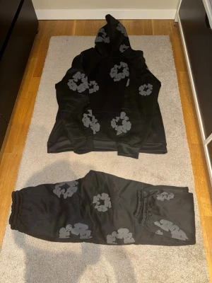 Svart hoodie från Denim Tears med blommor - Svart hoodie från Denim Tears med gråa blommönster tryckta över hela plagget. Hoodien har en klassisk passform med huva och långa ärmar. Perfekt för dig som vill sticka ut med en unik och trendig stil.
