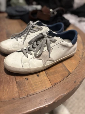 Golden Goose 41 - Stilrena vita sneakers från Golden Goose med grå snörning, beige stjärndetalj på sidan och mörkblå hälkappa. Skorna har en klassisk låg profil och är tillverkade i skinn med detaljer i mocka. Perfekta för en avslappnad och trendig look.