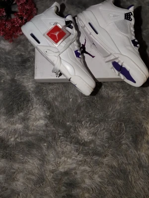 Nike Air Jordan 4 vit/lila sneakers - Säljer ett par Nike Air Jordan 4 sneakers i vitt med lila detaljer. Skorna har snörning, meshpaneler och klassisk rund tå. Sulan är vit med lila inslag och Jumpman-logga syns på plösen och undersidan. Perfekta för dig som vill sticka ut med stilrena och ikoniska sneakers.