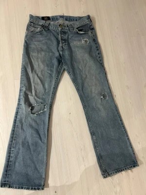 Lee jeans - Säljer ett par ljusblå bootcut jeans från Lee med slitna detaljer och hål på ena benet. Klassisk femficksmodell med Lee-lapp bak. Jeansen har en avslappnad passform och är tillverkade i bomull.