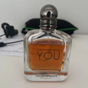 Emporio Armani Stronger With You parfym - Emporio Armani Stronger With You är en elegant parfym i en genomskinlig glasflaska med rundade former och silverfärgad metallkork. Doften har en varm bärnstensfärgad vätska och flaskan har stilren svart text på framsidan.