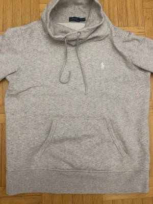 Grå hoodie från Polo Ralph Lauren - Snygg ljusgrå hoodie från Polo Ralph Lauren med klassisk huva och dragsko. Har en stor magficka och den ikoniska vita logon broderad på bröstet. Tillverkad i mjuk bomullsblandning som känns skön mot huden. Perfekt för en avslappnad och stilren look.