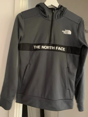 Mörkgrå huvtröja från The North Face - Mörkgrå hoodie med halv dragkedja och huva från The North Face. Vit logga på bröstet och ryggen samt svart band med vit text över bröstet. Tillverkad i mjukt funktionsmaterial och har långa ärmar.