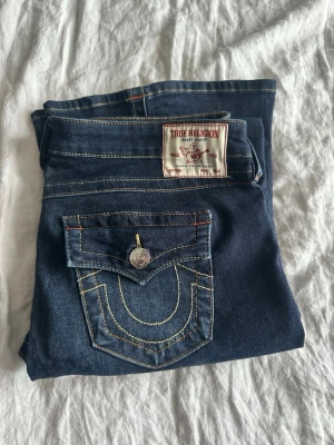 True Religion Joey Low Rise Flare Jeans - Mörkblå jeans från True Religion, modell Joey Low Rise Flare. Snygga kontrastsömmar och ikonisk fickdetalj med knappstängning bak. Klassisk patch på bakfickan och låg midja för en cool look. Perfekta för dig som gillar unik design och markerade detaljer. Aldrig använda, endast testade. 