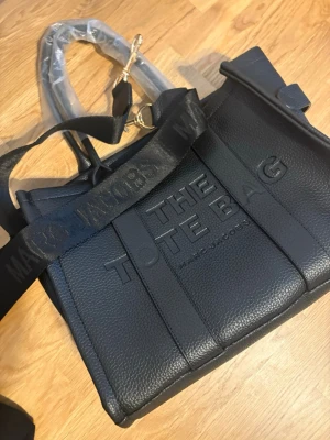 Svart The Tote Bag liknande  - Säljer en svart  väska med präglad logga framtill. , dragkedja upptill och rymligt innerfack. Perfekt för dig som vill ha en stilren och praktisk väska.