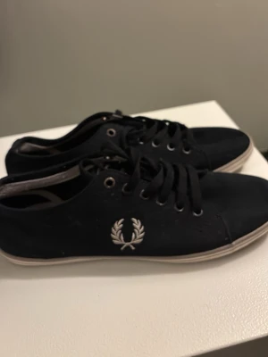 Svarta Fred Perry sneakers med logga 39 - Snygga svarta sneakers från Fred Perry med klassisk broderad logga i vitt på sidan. Skorna har snörning, vit sula och brunt rutmönster undertill. 