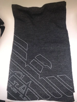 Mörkgrå halsduk från Emporio Armani - Snygg mörkgrå halsduk från Emporio Armani med stilrent mönster och EA-logga. Halsduken är stickad i mjukt material och har ett diskret, modernt uttryck med grafiska linjer och märkets ikoniska örn. Perfekt accessoar för att lyfta din outfit.