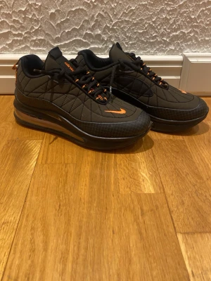 Svarta Nike Air Max sneakers med orange detaljer - Säljer ett par svarta Nike Air Max sneakers med orange detaljer och snörning. Skorna har en quiltad ovandel, rund tå och synlig Air Max-sula. Swoosh-loggan och detaljer på plösen är i orange, vilket ger en snygg kontrast till den mörka basen.