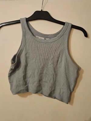 Ljusgrå/blå ribbad croppad linnetopp - Säljer en ljusgrå/blå croppad linnetopp med ribbad struktur. Toppen är ärmlös och har breda axelband samt rund halsringning. Perfekt att styla med höga jeans eller kjol för en trendig look.