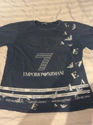 Blå Emporio Armani t-shirt XL - Snygg blå t-shirt från Emporio Armani med stora vita logotyper och tryck över hela framsidan. T-shirten har rund hals och korta ärmar. Perfekt för dig som gillar streetwear och vill sticka ut med ett exklusivt märke.