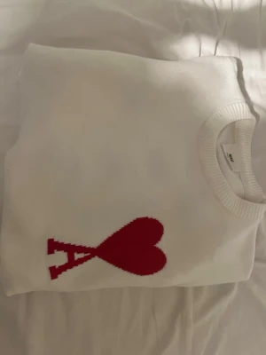 Vit stickad tröja från Ami Paris - Vit stickad tröja från Ami Paris med ikoniskt rött hjärta och bokstaven 'A' på bröstet. Tröjan har rund halsringning och ribbstickade muddar. Perfekt för dig som gillar stilrena och trendiga plagg med en twist. Aldrig andnvänd.