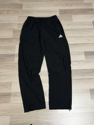 Svarta träningsbyxor från Adidas - Säljer ett par svarta träningsbyxor från Adidas med resår i midjan och dragkedjor vid bensluten. Byxorna har Adidas-loggan i vitt på vänster lår och är tillverkade i ett lätt och smidigt material som passar perfekt till träning eller sport.