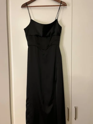 Svart satinklänning med smala axelband - Elegant svart klänning i satin med smala axelband och figurnära passform. Klänningen har en enkel och stilren design med lätt markerad midja och lång siluett. Perfekt för dig som gillar minimalistisk och tidlös stil. Klänningen är i storlek Xs-S och enbart använd en gång!