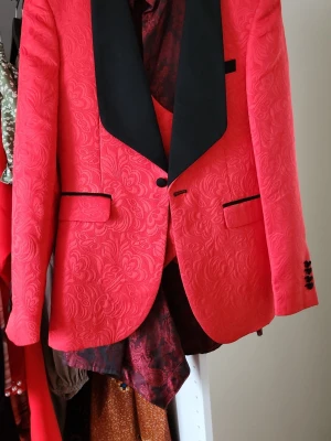 Jieweisi Röd kavaj och väst - Snygg röd kavaj och matchande väst med elegant blommigt mönster och svarta detaljer. Kavajen har svarta slag, fickor med svarta kanter och dekorativa knappar vid ärmslut. Västen har dubbla knapprader och samma blommiga struktur. Perfekt för dig som vill sticka ut.