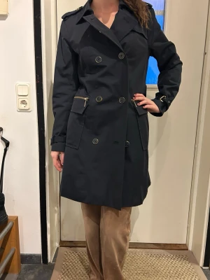 Michael kors- Svart trenchcoat med bälte och knappar - Klassisk svart trenchcoat med dubbelknäppning, axelklaffar och bälte i midjan. Jackan är lätt vadderad, fickor med lock och dekorativa detaljer i metall. Modellen är 175cm och 34/36