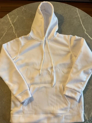 Vit hoodie med magficka och huva - Vit hoodie i storlek M, helt oanvänd, tillverkad i 100 % polyester som är både jätte skön och varm. Den mjuka insidan ger hög komfort, medan magfickan och snören i huvan ger en klassisk och praktisk design. Perfekt för både vardag och mysiga dagar – en hoodie du gärna använder om och om igen.👌