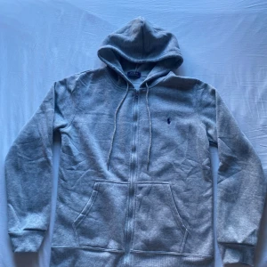 Grå hoodie från Polo Ralph Lauren - Snygg grå hoodie från Polo Ralph Lauren med dragkedja framtill, två stora fickor och justerbar huva med snören. Klassisk liten logga broderad på bröstet. Tillverkad i mjukt och bekvämt material, perfekt för en avslappnad stil.