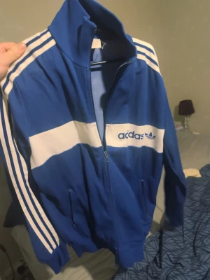 Vintage Adidas Originals track jacket – Made in Yugoslavia – M/L - Klassisk Adidas track jacket i blått och vitt med tre ränder och broderad trefoil-logga. Jackan är tillverkad i dåvarande Jugoslavien, vilket placerar den i slutet av 70-talet eller 80-talet – en period som många samlare och vintageintresserade uppskattar extra mycket. “Made in Yugoslavia” är ett tydligt kvalitets- och tidsmärke. Under den här eran producerades Adidas-plagg i mindre upplagor i Europa med fokus på slitstarka material, bra passform och gediget hantverk.