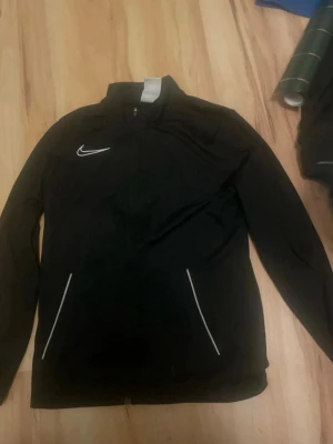 Svart Nike träningsjacka - Svart träningsjacka från Nike med vit logga på bröstet och vita detaljer vid fickorna. Jackan har hel dragkedja, hög krage och långa ärmar. Perfekt för sportiga aktiviteter och en clean look.