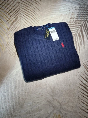 Mörkblå kabelstickad tröja Polo Ralph Lauren - Kabelstickad mörkblå tröja från Polo Ralph Lauren med klassisk röd broderad logga på bröstet. Rund halsringning och ribbade muddar. Perfekt för dig som vill ha en stilren och tidlös look.