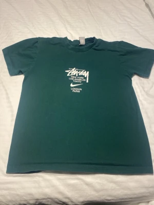 Grön Stüssy t-shirt med tryck - Mörkgrön t-shirt från Stüssy med vitt tryck på bröstet där det står Stüssy och flera storstäder. Klassisk passform och rund hals. Perfekt för dig som gillar streetwear och vill ha en enkel men snygg tee.