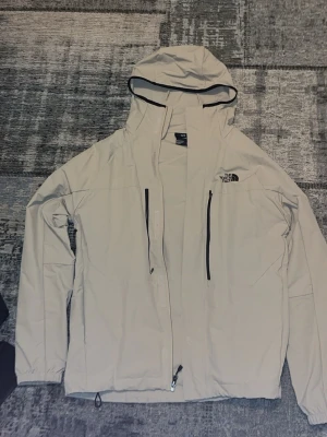 Beige vindjacka från The North Face - Snygg beige vindjacka från The North Face med huva och svarta dragkedjor framtill. Jackan har två vertikala bröstfickor och justerbar nederkant. Perfekt för blåsiga dagar och enkel att matcha med olika outfits.