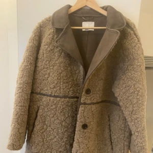 Beige teddyjacka med stora knappar - Mysig beige teddyjacka med stora knappar framtill och bred krage i kontrasterande material. Jackan har en loose passform och markerade sömmar som ger en cool vintagekänsla. Perfekt för dig som vill ha en varm och trendig jacka till hösten eller vintern.