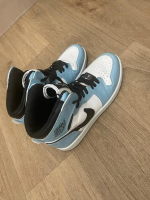Nike Air Jordan 1 blå och vita sneakers - Säljer ett par Nike Air Jordan 1 sneakers i blått och vitt med svarta detaljer och snörning. Skorna har hög siluett, svart swoosh och klassisk Air Jordan-logga på sidan. Perfekta för dig som vill ha en ikonisk och stilren sneaker.