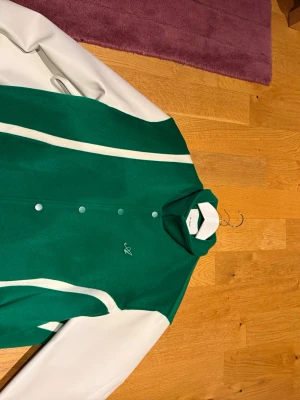 Grön och vit overshirt  Axel Arigato - Snygg grön och vit overshirt från Axel Arigato med vita detaljer och tryckknappar framtill. Jackan har klassisk college-look med kontrasterande ärmar och diskret broderad logga på bröstet. Perfekt för dig som vill sticka ut med stilren design. Priset går att diskutera vid intresse 