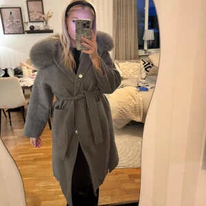 Grå kappa med - Snygg grå kappa med päls på kragen och knytbälte i midjan. Kappan har långa ärmar, knappar framtill och en klassisk, rak passform. Perfekt för kyliga dagar när du vill vara både varm och stilren. Den är i ull så är väldigt varm 