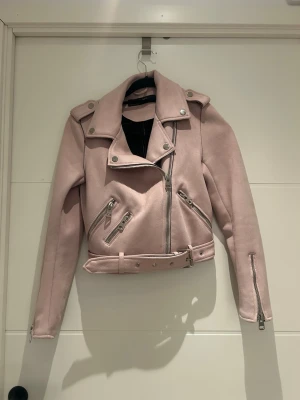 Ljusrosa skinnjacka med bälte - Snygg ljusrosa skinnjacka i bikerstil med flera dragkedjor, bred krage och bälte i midjan. Jackan har långa ärmar och coola detaljer i silver. Perfekt för dig som vill sticka ut med en trendig look.