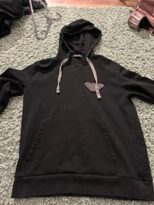 Svart hoodie från Emporio Armani - Svart hoodie från Emporio Armani med grå dragsnören och broderad logga på bröstet. Klassisk känguruficka framtill och mjukt material som känns skönt mot huden. Perfekt för en chill och stilren look.