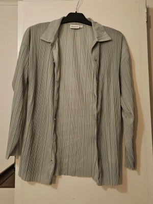 Grön plisserad skjorta  - Säljer en grå plisserad skjorta från XS/S. Skjortan har lång ärm, klassisk krage och knappar framtill. Materialet är lätt och har en snygg struktur med tunna vertikala veck som ger en modern vibe.