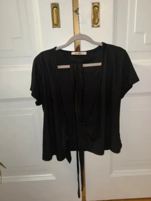 Svart kortärmad kofta från Nakd - Kortärmad svart kofta från Nakd med öppen front och långa band som kan knytas framtill. Tunn och mjuk trikå som faller snyggt. Perfekt att slänga över ett linne för en avslappnad look.