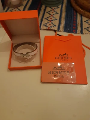 Hermès vit och silver armband - Snyggt armband från Hermès i vit emalj med silverfärgade metalldetaljer och klassiskt H-spänne. Kommer i original orange ask och påse från Hermès. Perfekt accessoar för dig som gillar exklusiv och stilren design.