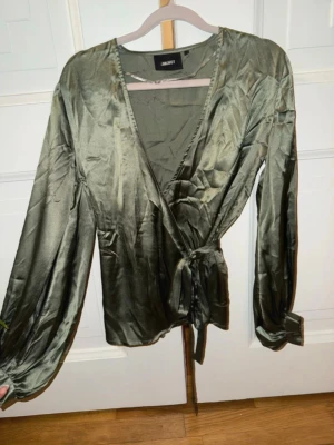 Olivgrön omlottblus från Object - Snygg olivgrön omlottblus från Object i glansigt satinliknande material. Blusen har långa ärmar med breda manschetter och knytband i midjan för en feminin siluett. Känsligt material därav har några små masker gått upp. Syns inte tydligt, men vill ändå nämna det. 